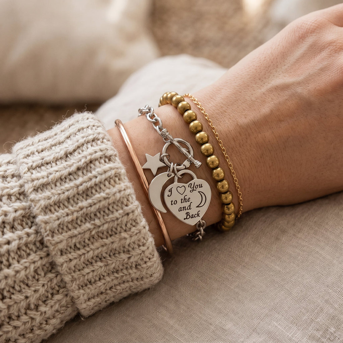 A wrist wearing a stack of mixed-metal bracelets over a neutral knit sweater sleeve